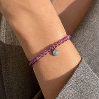 Close-up of a wrist wearing a purple beaded bracelet with a silver charm on a textured surface.