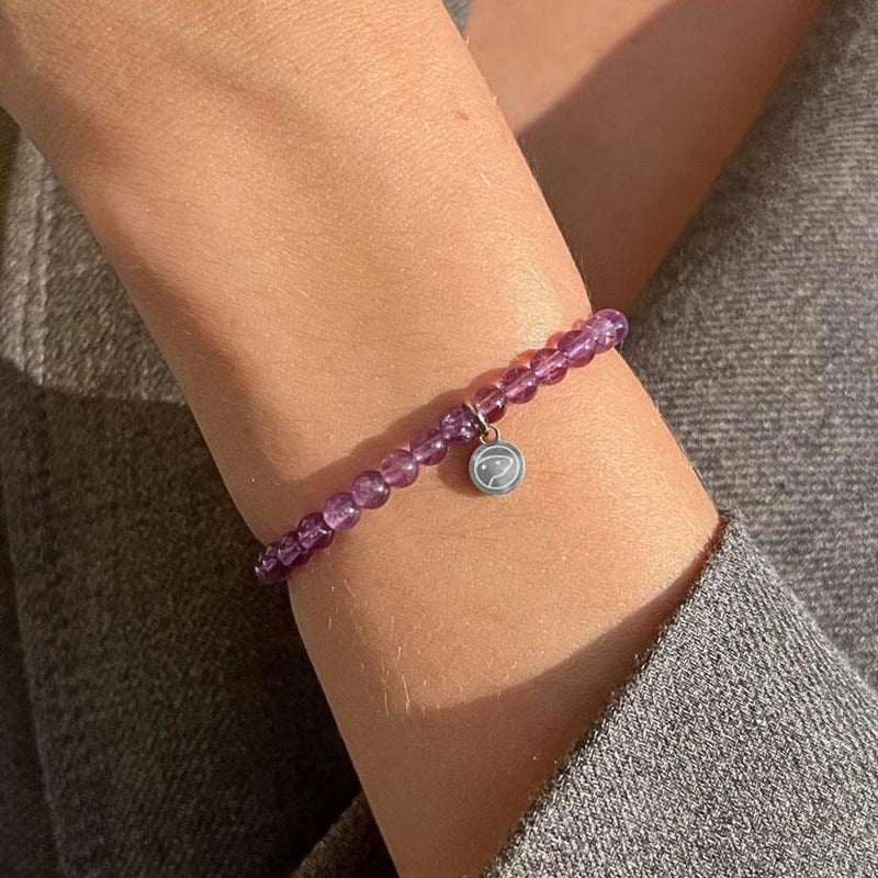 Close-up of a wrist wearing a purple beaded bracelet with a silver charm on a textured surface.