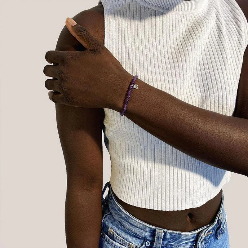 Person wearing a white ribbed tank top and blue jeans with a purple bracelet on a plain background