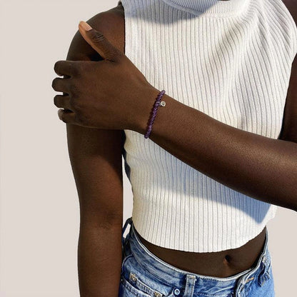 Person wearing a white ribbed tank top and blue jeans with a purple bracelet on a plain background