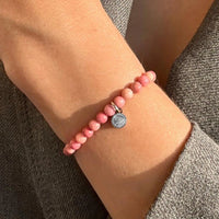 Pink beaded bracelet with a gray charm on a wrist against a neutral background